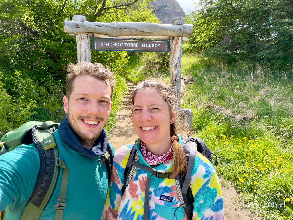 Two hikers at the Fitz Roy trailhead in El Chaltén, Argentina, standing beneath the Senderos Torre | Fitz Roy entrance sign before starting the hike.