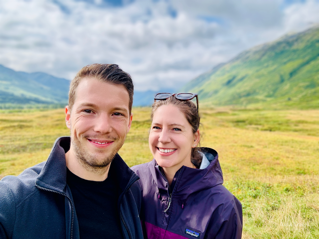 Smiling travel couple standing in front of a scenic mountain valley in Alaska.