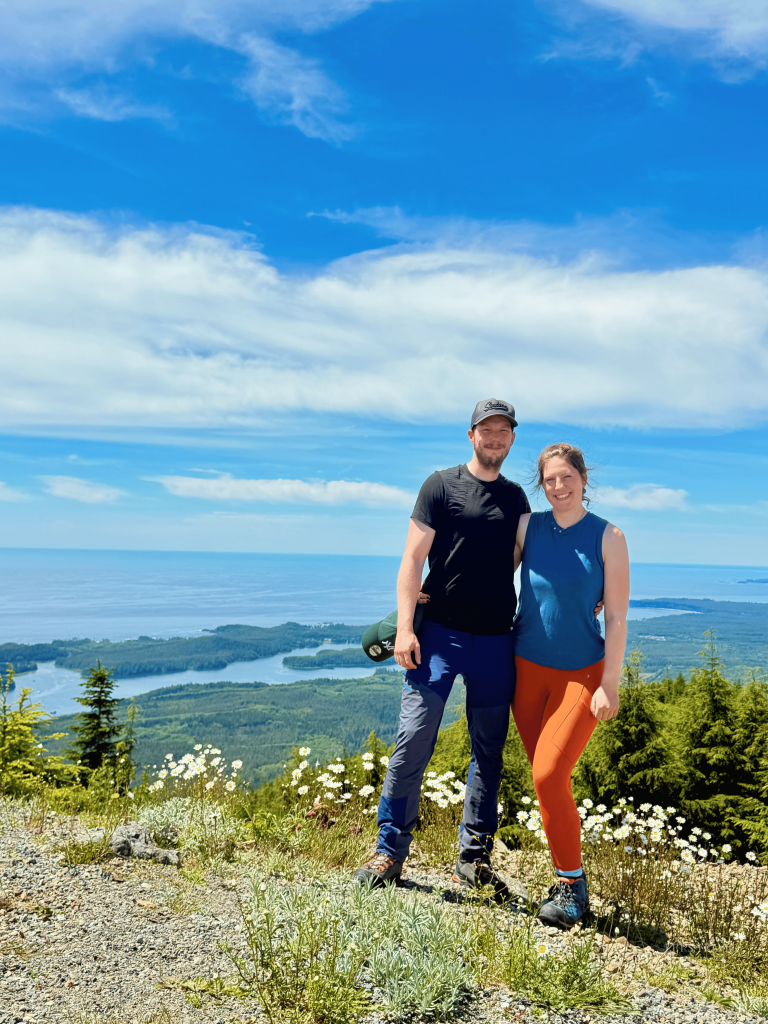 Mount Ozzard Hike on Vancouver Island with views of the surrounding coastline. Couple standing at a Mount Ozzard viewpoint on Vancouver Island, overlooking forested hills, coastal inlets and the Pacific Ocean.